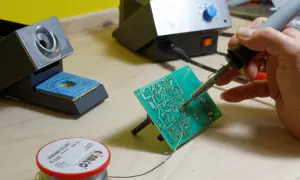 Ręka trzyma lutownicę do elektroniki, lutując zieloną płytkę drukowaną. Obok stojak na lutownicę i szpulka cyny.