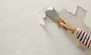 Dłoń z kielnią usuwa łuszczący się tynk ze ściany. Szybko usunąć stary tynk to pierwszy krok do remontu.