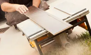 Ręce przycinają panel podłogowy na pile stołowej. Tarcze do cięcia paneli podłogowych pracują, tworząc pył.