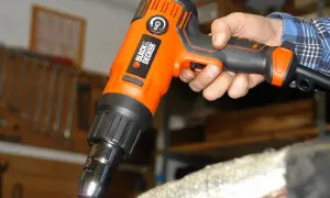 Pomarańczowa opalarka Black & Decker w dłoni, gotowa do pracy. W tle warsztat z narzędziami.