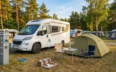 Na kempingu kamper podłączony do prądu. Na słupku informacja: NAPIĘCIE 230V. Obok namiot i meble kempingowe.