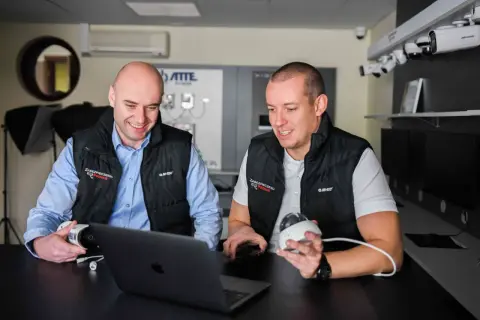 Dwóch mężczyzn omawia systemy monitoringu. Jeden trzyma kamerę, drugi patrzy na laptopa, planując zdalny monitoring.