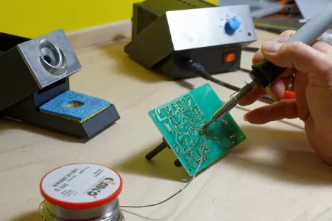 Ręka trzyma lutownicę do elektroniki, lutując zieloną płytkę drukowaną. Obok stojak na lutownicę i szpulka cyny.