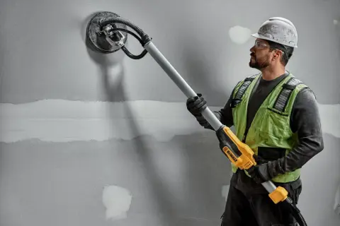 Budowlaniec szlifuje ścianę za pomocą szlifierki do gipsu DeWalt.