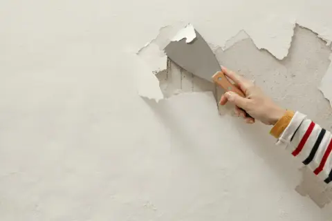 Dłoń z kielnią usuwa łuszczący się tynk ze ściany. Szybko usunąć stary tynk to pierwszy krok do remontu.