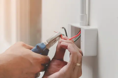 Elektryk podłącza przewody, używając szczypiec. Widoczne oznaczenie prądu L na czerwonym kablu.