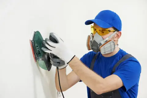 Pracownik w masce i okularach ochronnych wykonuje ręczne szlifowanie płytek na białej ścianie.