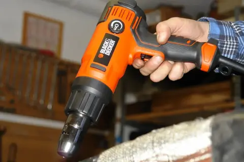 Pomarańczowa opalarka Black & Decker w dłoni, gotowa do pracy. W tle warsztat z narzędziami.