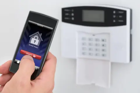 Dłoń trzyma smartfon z aplikacją do ochrony mieszkania w bloku. Na ekranie widnieje przycisk "Disarm". Obok panel alarmowy.