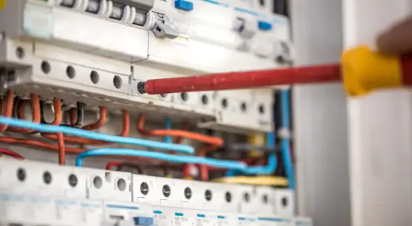 Ręka z czerwonym śrubokrętem pracuje przy panelu z wyłącznikami i kolorowymi przewodami. To instalacja niskoprądowa.