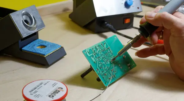 Ręka trzyma lutownicę do elektroniki, lutując zieloną płytkę drukowaną. Obok stojak na lutownicę i szpulka cyny.