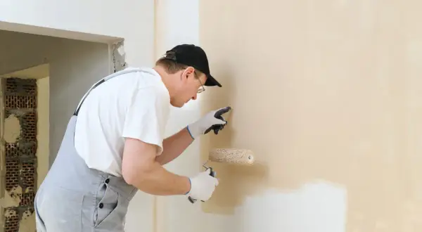 Mężczyzna w ogrodniczkach i czapce maluje ścianę wałkiem. Lampa do malowania ścian jest niezbędna do precyzyjnego wykończenia.