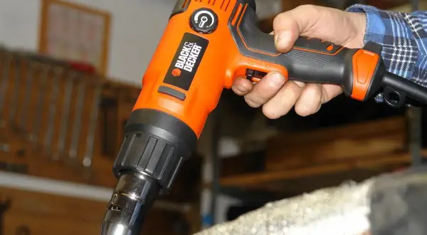 Pomarańczowa opalarka Black & Decker w dłoni, gotowa do pracy. W tle warsztat z narzędziami.