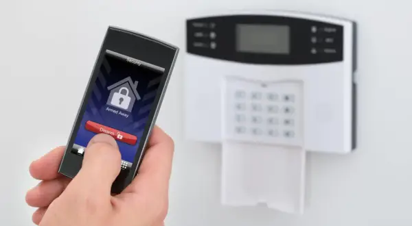 Dłoń trzyma smartfon z aplikacją do ochrony mieszkania w bloku. Na ekranie widnieje przycisk "Disarm". Obok panel alarmowy.