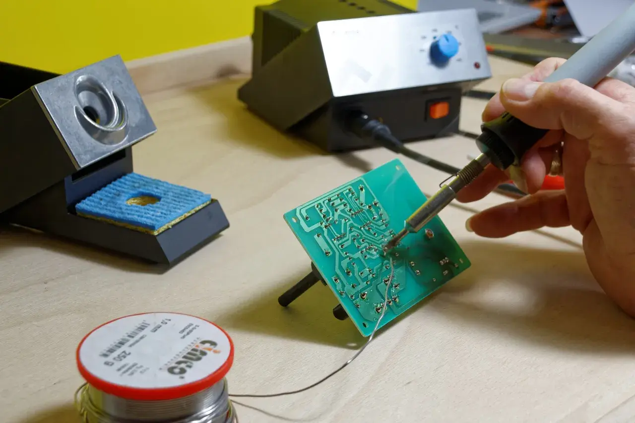 Ręka trzyma lutownicę do elektroniki, lutując zieloną płytkę drukowaną. Obok stojak na lutownicę i szpulka cyny.