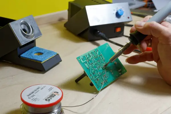 Ręka trzyma lutownicę do elektroniki, lutując zieloną płytkę drukowaną. Obok stojak na lutownicę i szpulka cyny.